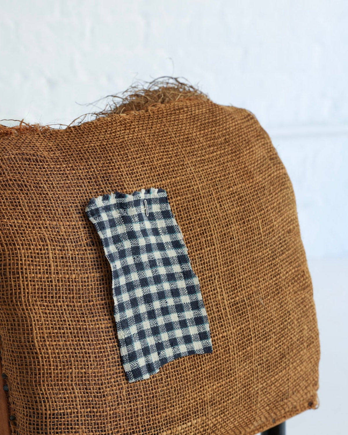 A close-up of a rustic brown burlap fabric draped over a chair, with a small navy and white gingham fabric patch attached to the front, set against a bright white wall and floor.