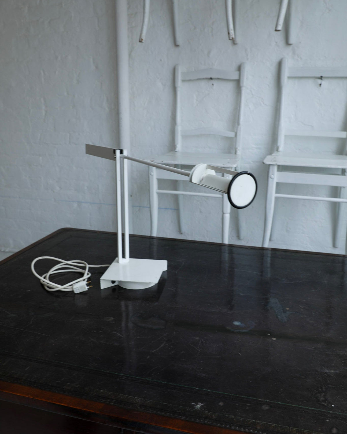 A modern white adjustable desk lamp with a circular shade and cord, placed on a dark wooden table in front of a white brick wall with white chairs.