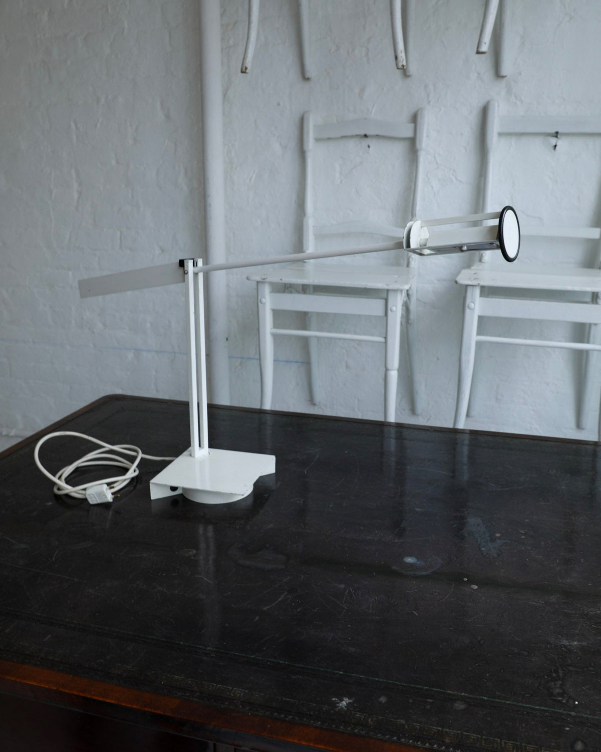 A modern white adjustable desk lamp with a rectangular base, slim arm, and cylindrical shade, placed on a dark wooden table in a room with white chairs against a white wall.