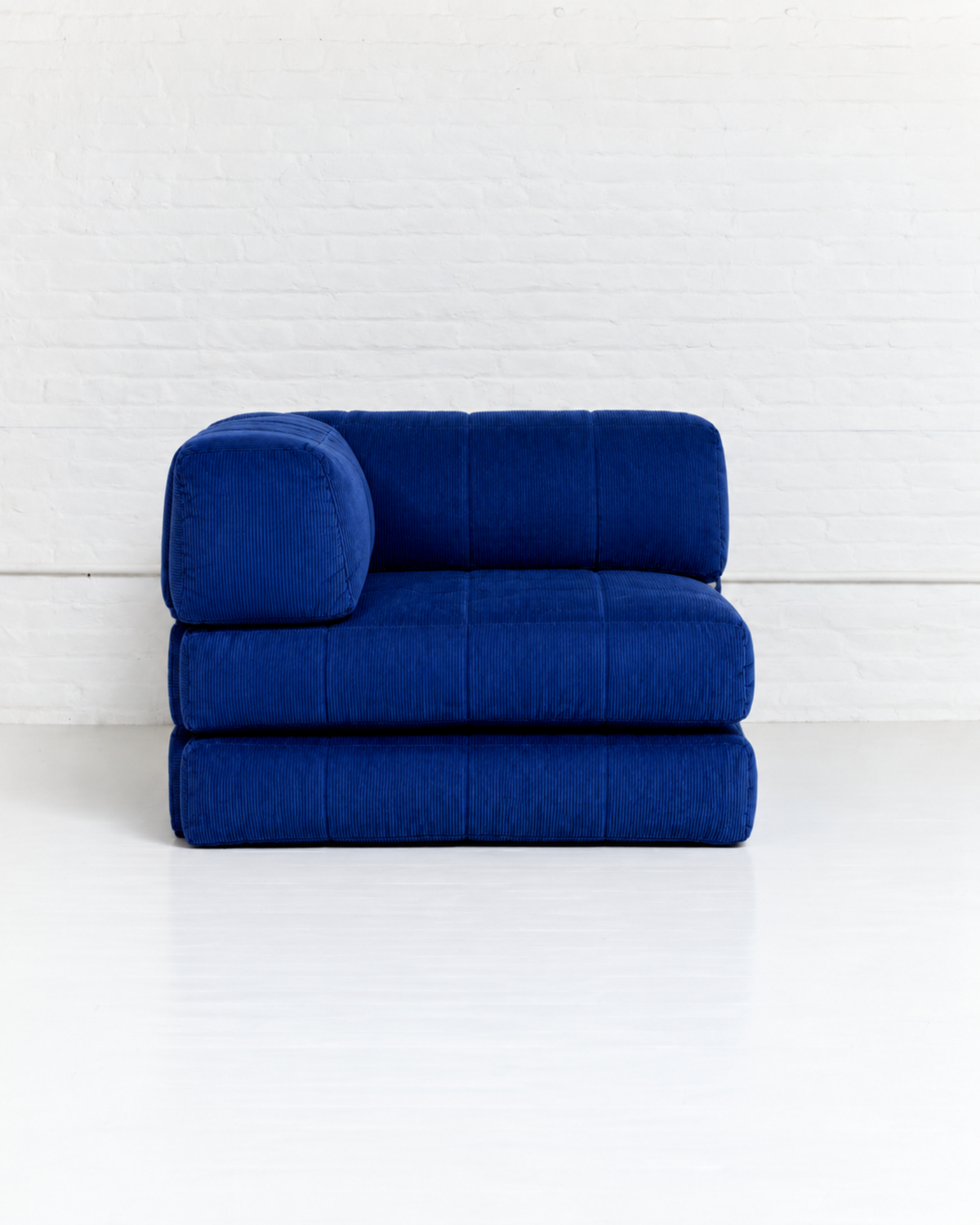 A modern modular floor cushion chair in deep royal blue velvet, featuring a thick segmented seat, a matching cylindrical backrest, and a low-profile base, set against a white brick wall and white floor.