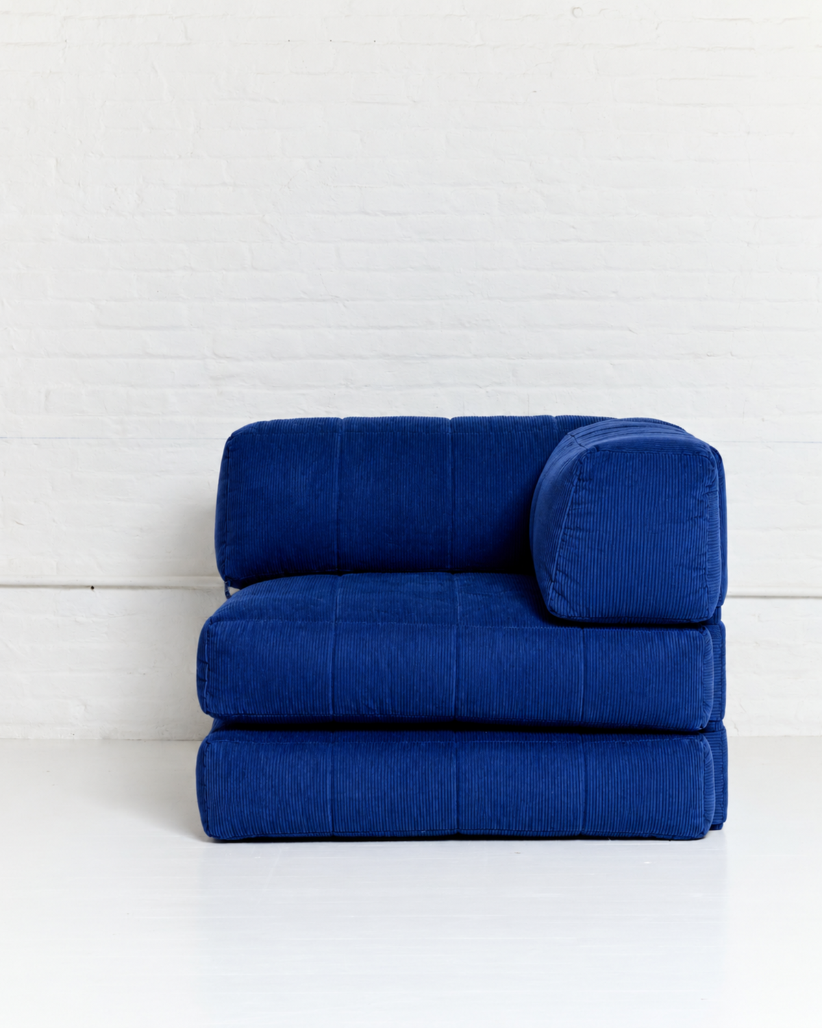 A modern modular floor cushion chair in a deep royal blue corduroy fabric, featuring a thick seat, a matching backrest, and a separate cylindrical lumbar pillow, set against a white brick wall.