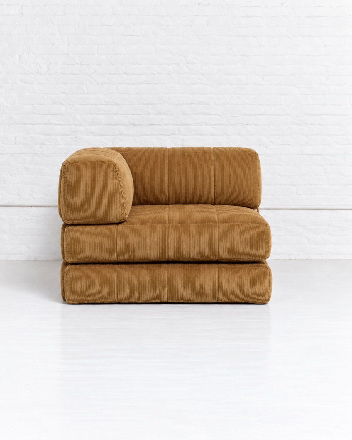 A modern modular upholstered ottoman or small sofa in a warm camel brown velvet fabric, set against a white brick wall and white floor.