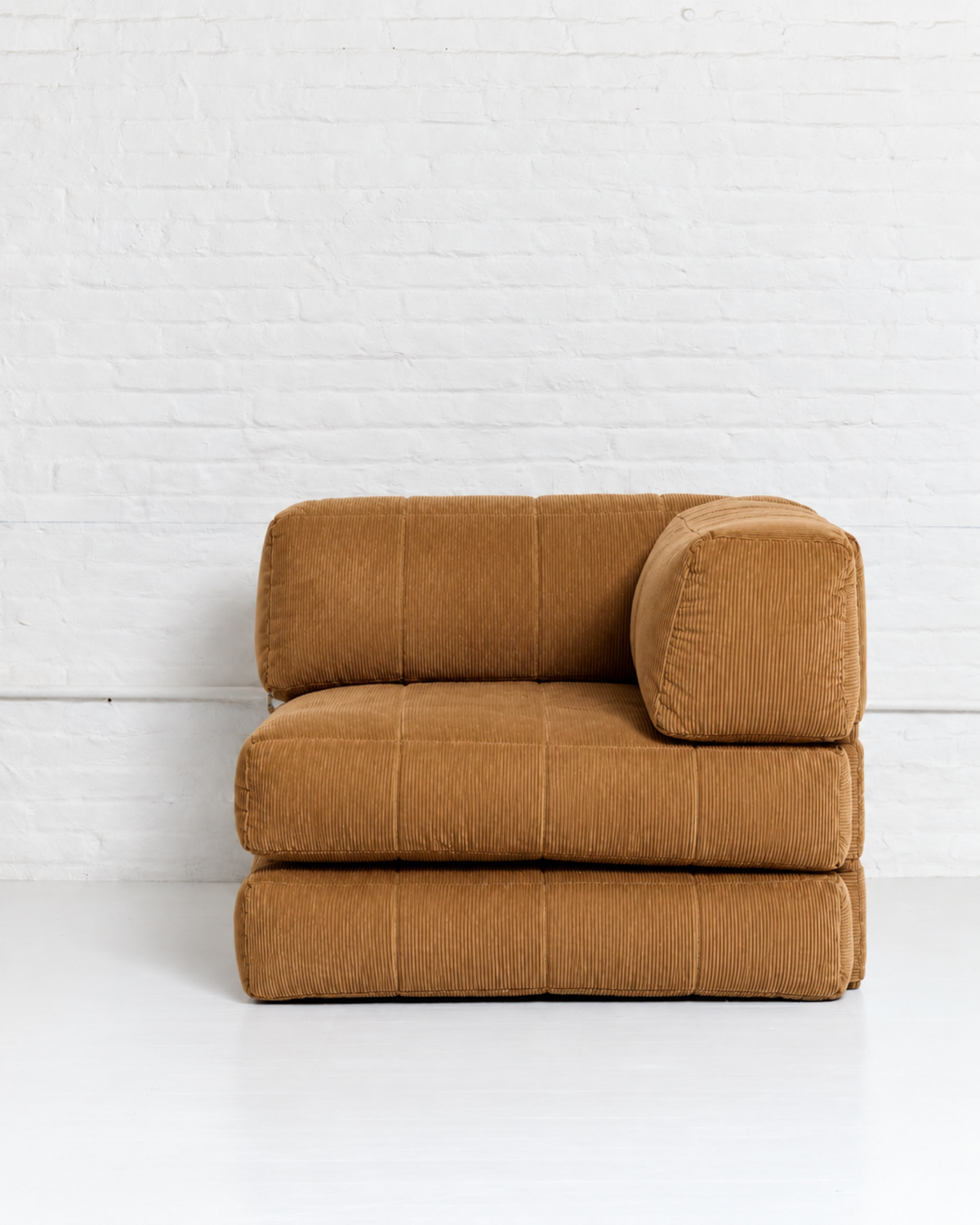 A low-profile, modular upholstered floor cushion or small chair in a warm camel brown corduroy fabric, set against a white brick wall and white floor.