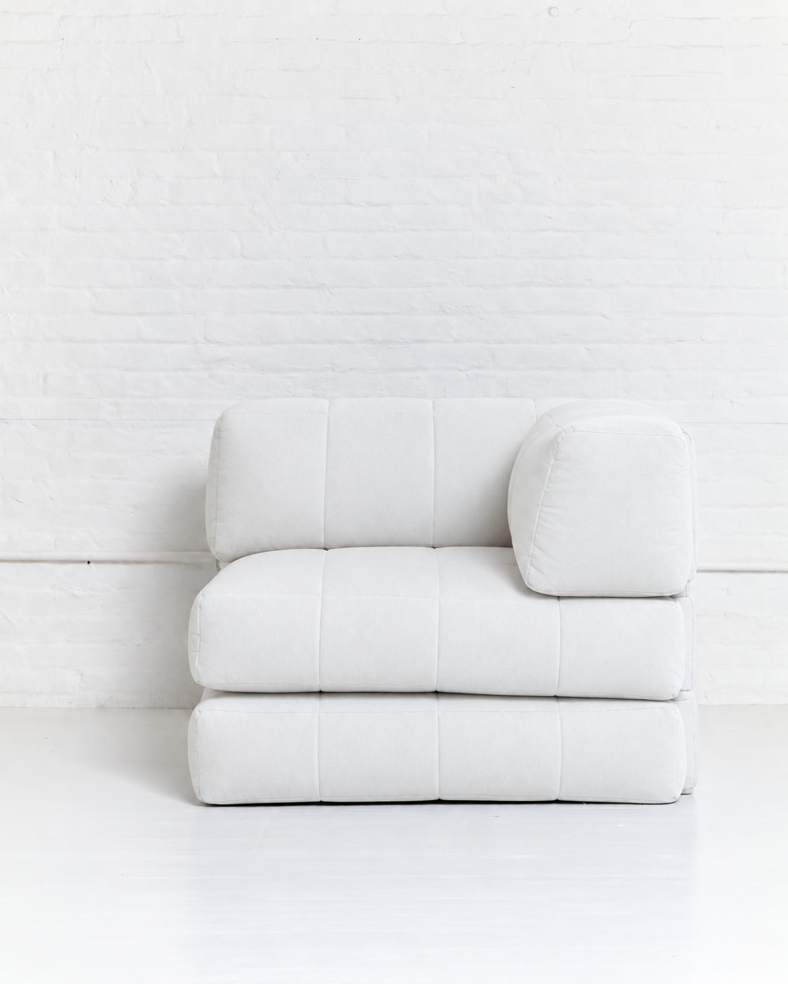 A modern modular white upholstered ottoman or low seat with a square backrest cushion and a separate cylindrical lumbar pillow, set against a white brick wall and white floor.
