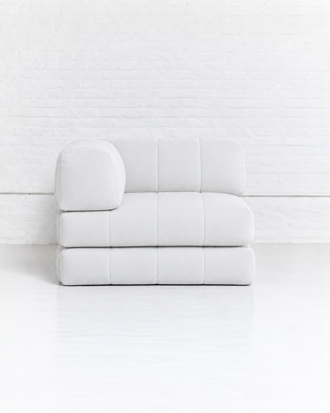 A modern, low-profile modular sofa upholstered in a very light gray or off-white fabric, featuring a tufted grid pattern and a rounded left armrest, set against a white brick wall and white floor.