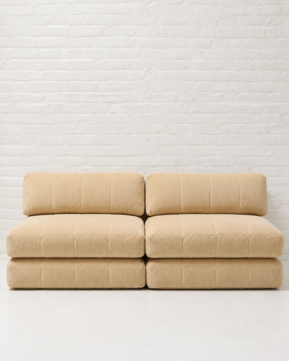 A modern modular upholstered seating unit made of four square cushioned blocks in a light beige fabric, set against a white brick wall background.