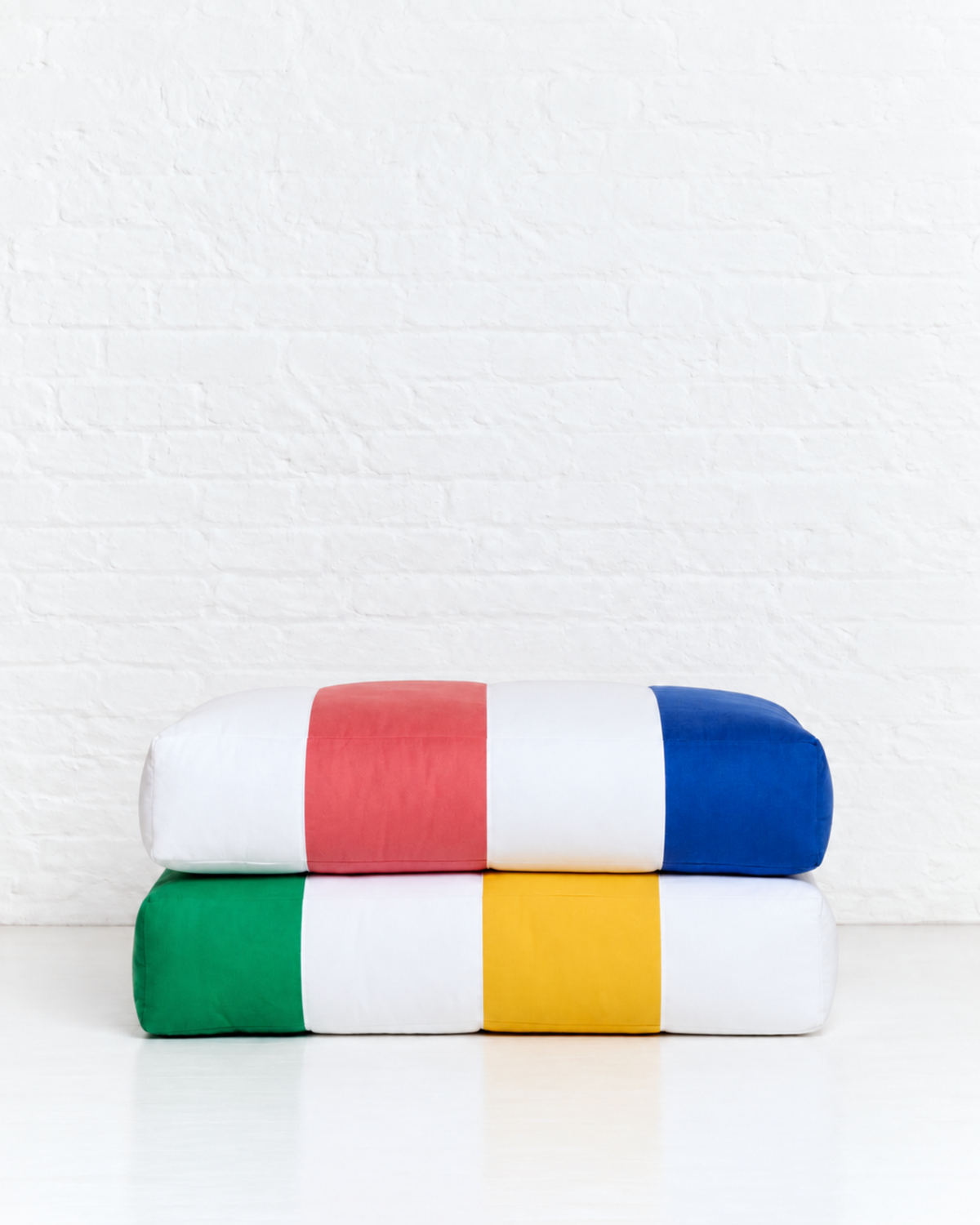 Two folded rectangular pillows stacked on a white surface against a white brick wall. The top pillow has vertical stripes in white, red, and blue, and the bottom pillow has vertical stripes in green, white, and yellow.