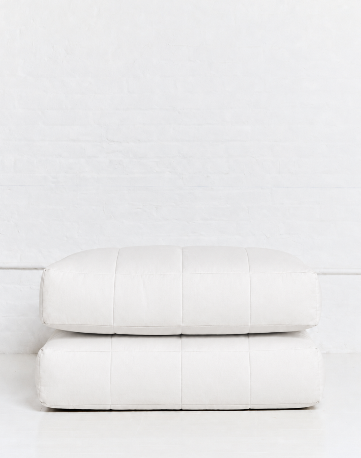 Two stacked rectangular white cushions or floor poufs with a quilted stitched pattern, set against a white brick wall and white floor.