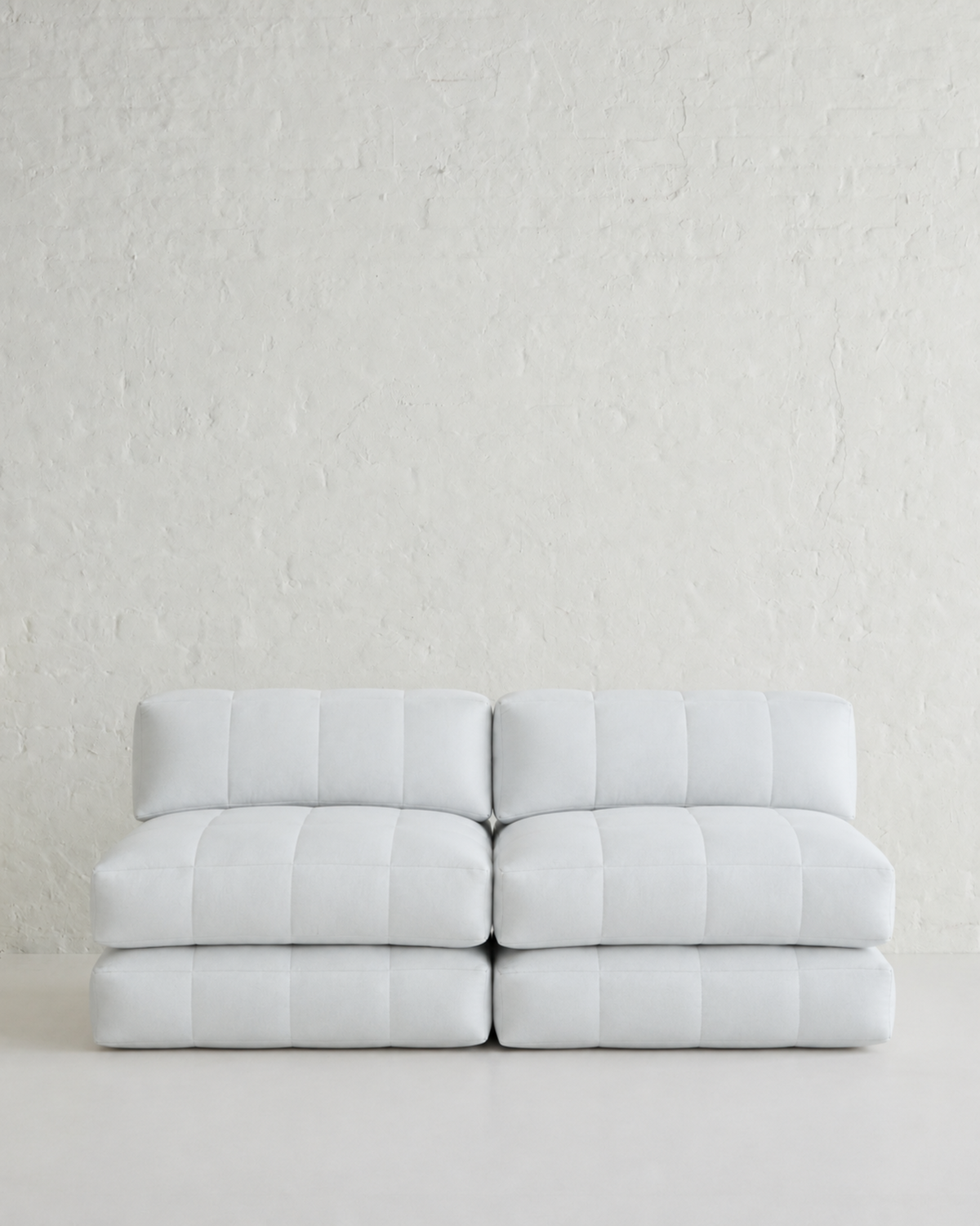 A modern two-seater modular sofa upholstered in white tufted leather, positioned against a textured white brick wall on a light gray floor.