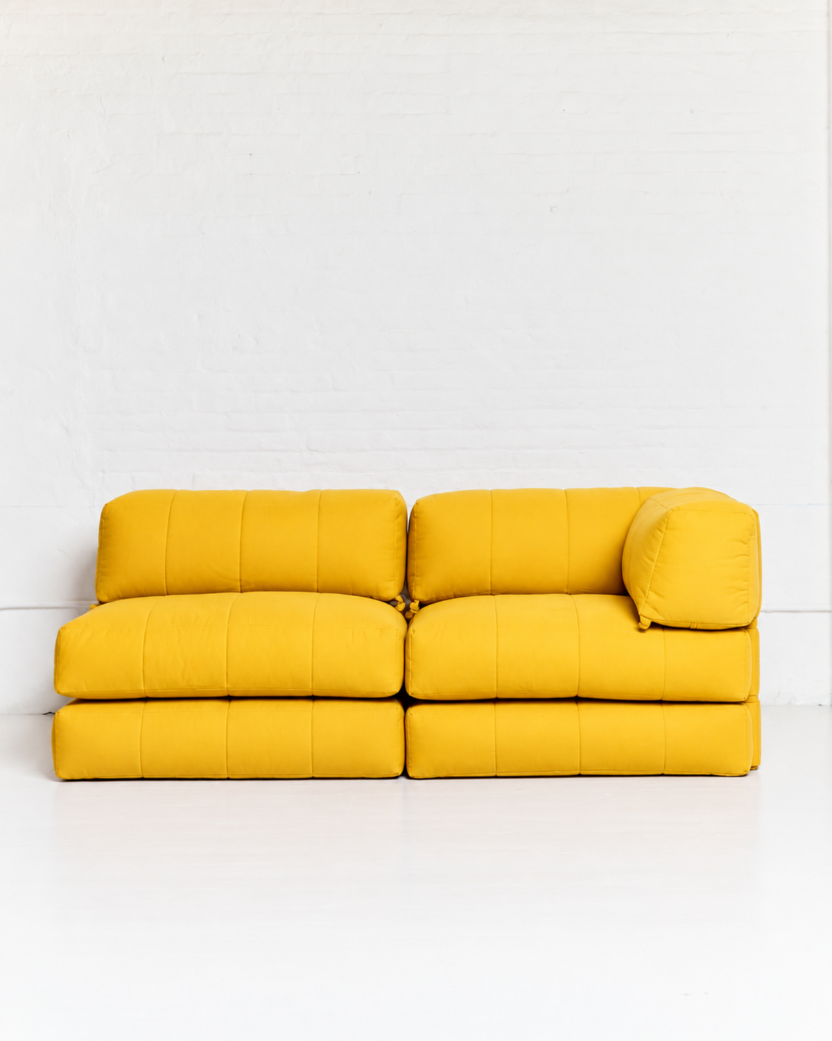 A modern modular yellow sofa with a low, blocky design and channel-tufted cushions, set against a white brick wall and white floor.